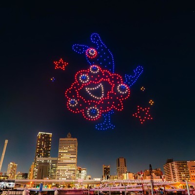"A drone light display over a city skyline at night, featuring a large, colorful character in red, blue, and white lights. The character has a rounded, flower-like shape with a smiling face and stars around it. The city buildings are illuminated below, giving a vibrant atmosphere to the scene." 
