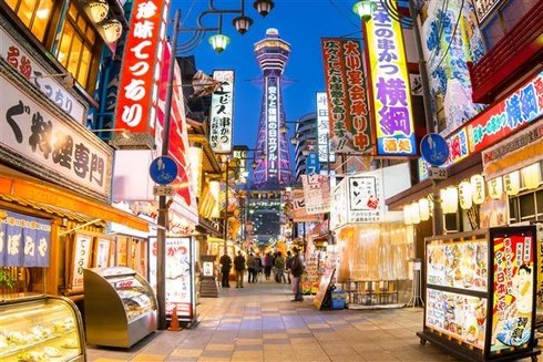 "A bustling street in Osaka's Shinsekai District, featuring bright neon signs and colorful storefronts under a twilight sky. The iconic Tsutenkaku Tower, lit up with purple and yellow lights, stands prominently in the background. Japanese signs for various shops, restaurants, and advertisements fill both sides of the street. People walk along the vibrant street lined with food stalls and restaurant displays 